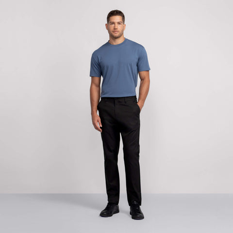 A man stands against a plain light gray background, wearing a blue short-sleeve t-shirt, black shoes, and Jack Archer Jetsetter Tech Pant Relaxed Fit in True Black with one hand in his pocket and a neutral expression.