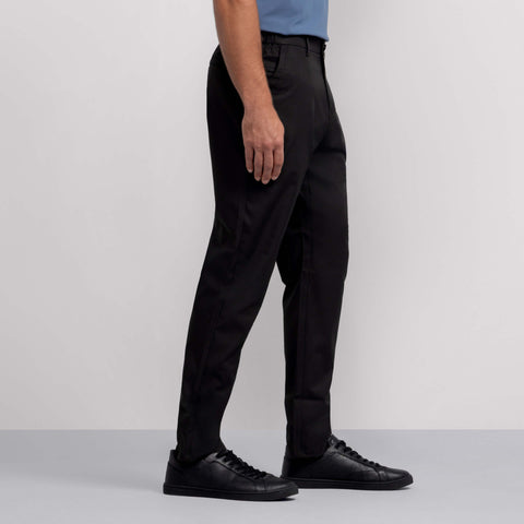 A person stands sideways against a plain light gray background, showing only the lower half of the body, wearing Jack Archer's Jetsetter Tech Pant Relaxed Fit in True Black with a blue shirt and black sneakers.