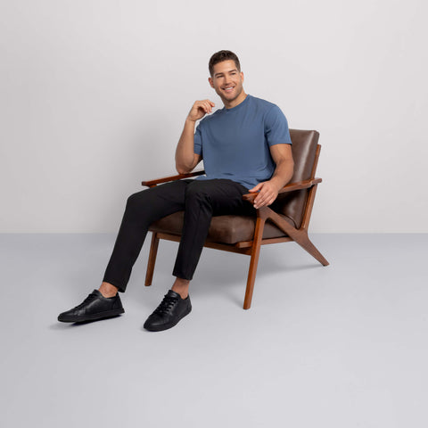 A man wearing a blue t-shirt, Jack Archer Jetsetter Tech Pant Relaxed Fit in True Black, and black shoes sits and smiles in a brown wooden chair against a plain light gray background.