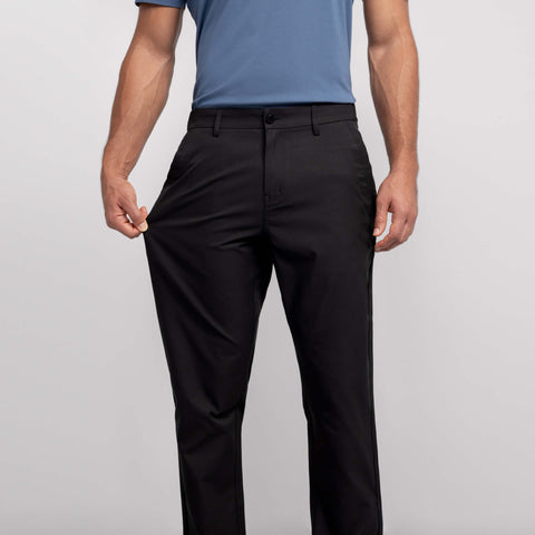 A man faces forward, pulling the side of his Jetsetter Tech Pant Relaxed Fit in True Black by Jack Archer to show extra room in the waistband and leg. He wears a blue polo; the photo is cropped at his shoulders and ankles.