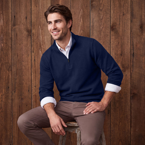 A man in a navy quarter-zip sweater and taupe pants seated against a wood plank wall.
