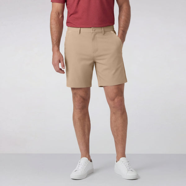 A man stands against a plain light background wearing a red short-sleeve shirt, white sneakers, and Jack Archer’s Jetsetter Tech Short 7" in Sand Tan, with one hand in his pocket.