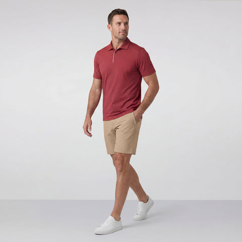 A man wearing Jack Archer's Jetsetter Tech Short 7" in Sand Tan, a red polo, and white sneakers stands against a plain light gray background, looking to the side with his hands in his pockets.