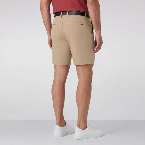 A man faces away, wearing a red shirt and Jack Archer's Jetsetter Tech Short 7" in Sand Tan with a black belt and white sneakers, set against a plain light background.