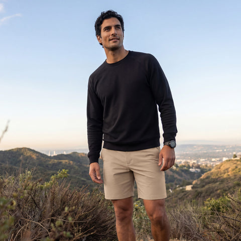 A man in a black long-sleeve shirt and Jack Archer Jetsetter Tech Short 7" in Sand Tan stands on a hillside trail with cityscape and mountains in the background beneath a clear sky.