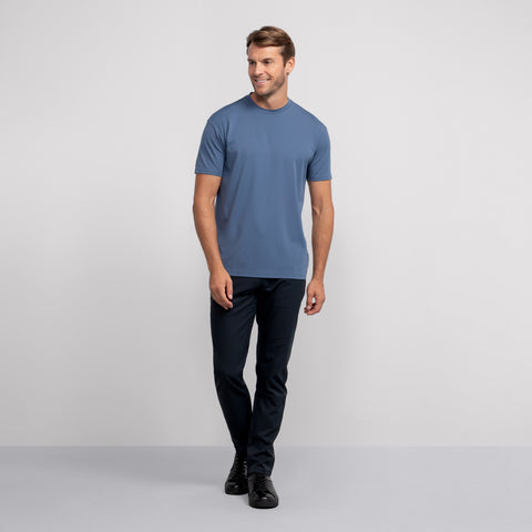 A man, smiling and walking leftward, wears the Jetsetter Tee—a breathable performance crewneck—paired with dark pants and black shoes against a plain light gray background.