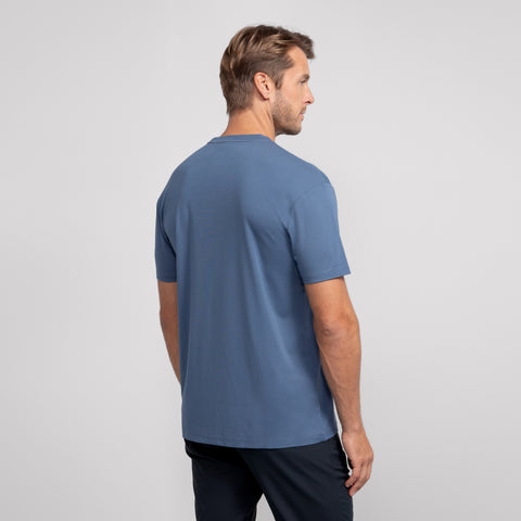 A man with short light brown hair is standing and facing away, wearing a blue Jetsetter Tee and dark pants against a plain light gray background.