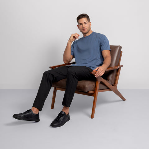 A man relaxes in a wooden armchair, wearing the Jetsetter Tee—a breathable blue crewneck—paired with black pants and sneakers, one hand on the armrest and the other on his face, set against a plain light background.