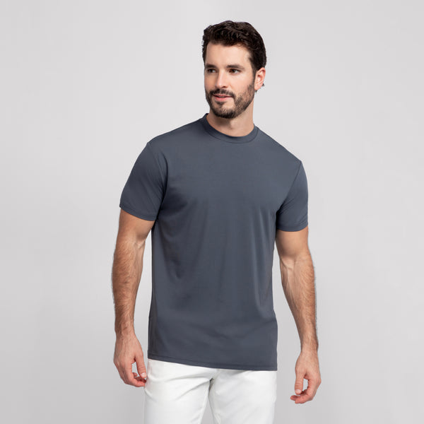A man with short dark hair and a beard stands against a plain light gray background, wearing the Jack Archer Jetsetter Tee in Charcoal Gray and white pants. He looks slightly left and smiles softly.