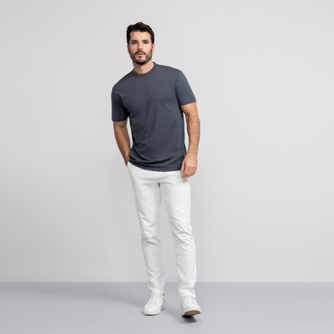A man stands against a plain light gray background, wearing the Jack Archer Jetsetter Tee in Charcoal Gray with white pants and sneakers. He looks straight ahead, one hand in his pocket.