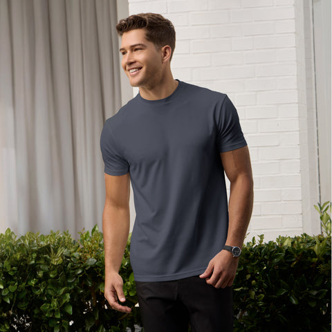 A smiling man stands outdoors in front of white curtains and a white brick wall, wearing the Jack Archer Jetsetter Tee in Charcoal Gray, black pants, and a watch, with green shrubs visible in the background.
