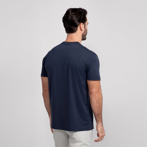 A man with short dark hair faces away, wearing the Jack Archer Jetsetter Tee in Deep Blue with a classic crewneck and light pants against a light gray background—ideal for any travel-ready wardrobe.
