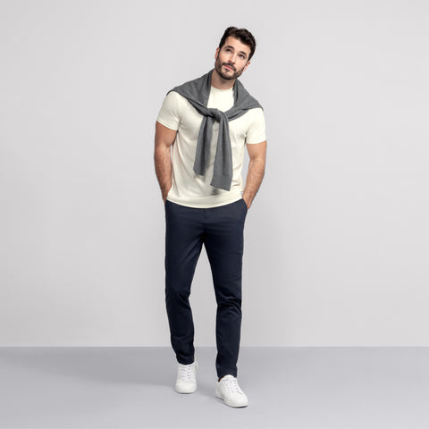 A man with short dark hair and a beard stands against a light gray background, hands in pockets, wearing the Jack Archer Jetsetter Tee in Ecru, navy pants, white sneakers, and a gray sweater over his shoulders for an easy travel look.