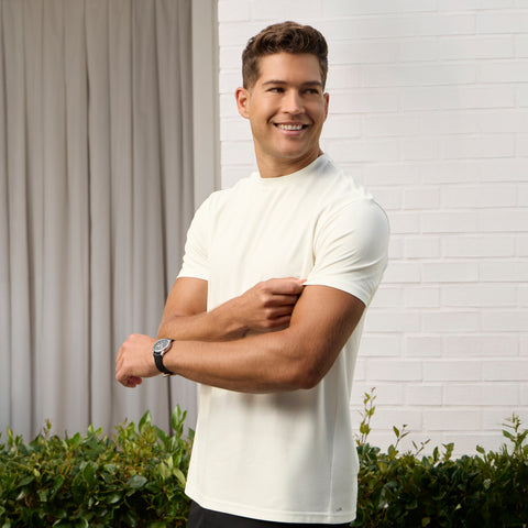 A man wears the Jack Archer Jetsetter Tee in Ecru and a wristwatch, standing outside by a white brick wall and greenery, smiling to his left with one arm bent across his chest.
