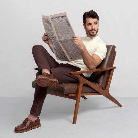 A man with dark hair and a beard, wearing the Jack Archer Jetsetter Tee in Ecru, brown pants, and loafers, sits cross-legged on a wooden armchair reading a newspaper against a plain light background.