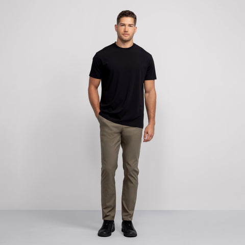 A man stands against a light gray background in the Jetsetter Tee and olive green pants. The breathable black crewneck pairs well with his black shoes as he gazes at the camera, one hand casually in his pocket.