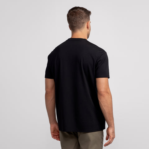 A man faces away from the camera in a Jack Archer Jetsetter Tee -- True Black and olive green pants against a light gray backdrop—an ideal travel-ready outfit.