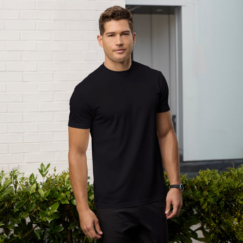 A man with short brown hair wears the Jack Archer Jetsetter Tee in True Black and black pants, standing outdoors before a white brick wall and green shrubs, looking directly at the camera with a neutral, travel-ready style.