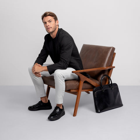 A man in a black blazer, Jack Archer Jetsetter Tee -- True Black, light gray pants, and black sneakers sits on a brown leather chair. His travel-ready black leather briefcase rests beside him against a plain, light background.