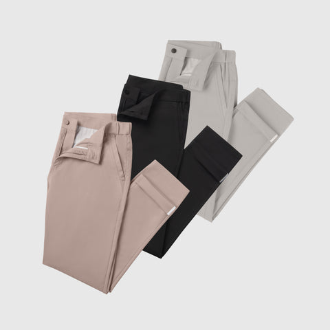 Three pairs of Jetsetter Tech Pant Slim Fit from the 3-Pack, in light pink, black, and light gray, are arranged diagonally on a white background. Each pair is neatly buttoned and partially unzipped.