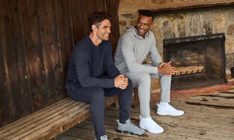 Two men in navy and heather grey sweatsuits seated on a porch bench beside a stone fireplace.