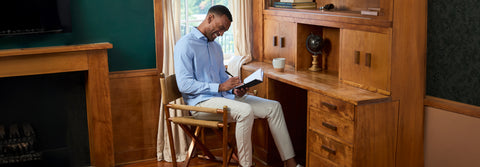 A man in a light blue shirt and stone trousers writing in a notebook at a wooden desk.