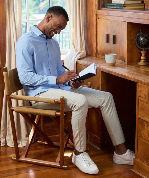 A man in a light blue shirt and stone trousers writing in a notebook at a wooden desk.
