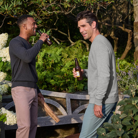 Two men holding beer bottles in a garden, one in a heather gray crewneck sweatshirt.