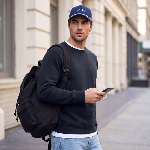 A man in a midnight navy crewneck sweatshirt, midnight navy cap and backpack on a city street.