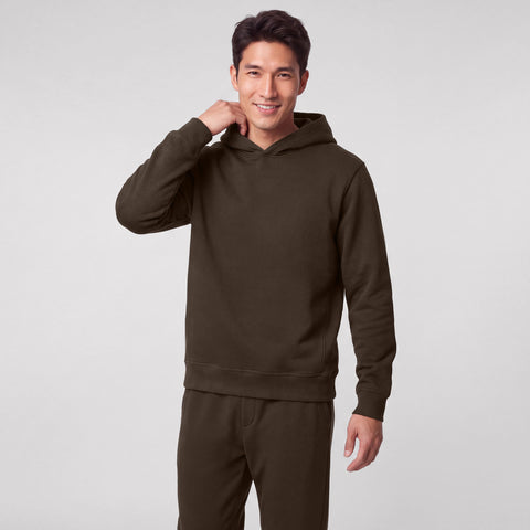 A man in an espresso brown hooded sweatshirt and matching joggers, smiling with one hand at his neck.