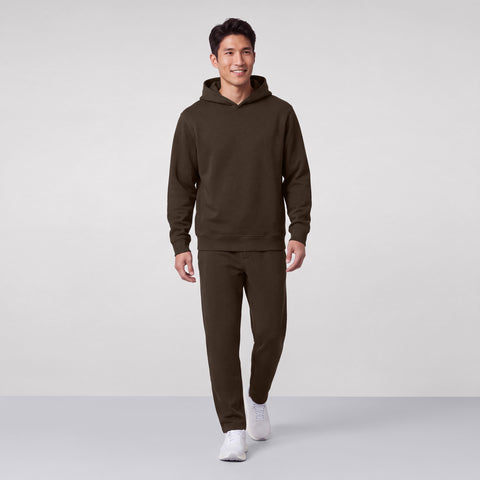 A man standing in an espresso brown hooded sweatshirt and matching joggers with white sneakers.
