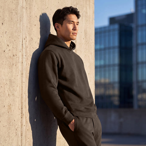 A man in an espresso brown hooded sweatshirt and joggers leaning against a concrete wall outdoors.