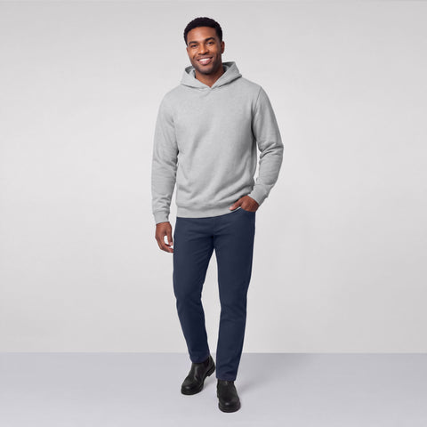 A man standing in a heather gray hooded sweatshirt, navy pants and black boots.