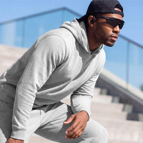A man in a heather gray hooded sweatshirt and joggers, wearing sunglasses and a cap, crouched on outdoor steps.