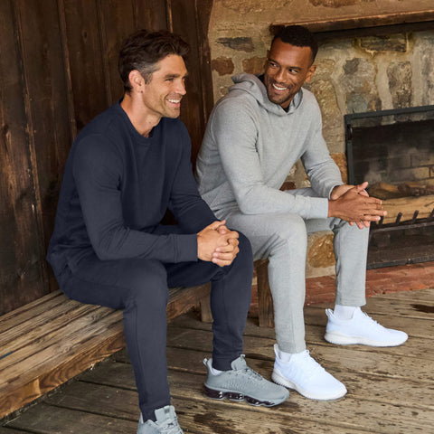Two men seated on a rustic bench, one in a heather gray hooded sweatshirt and joggers, the other in navy.