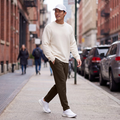 A man in espresso brown sweatpants and a cream sweater walking down a city sidewalk.