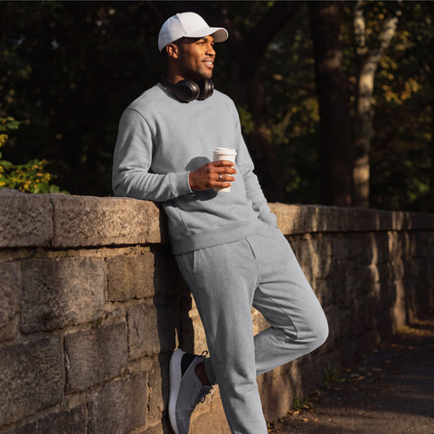 A man in a heather gray sweatshirt and matching sweatpants leaning on a stone wall with a coffee.