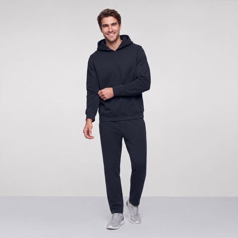 Smiling man in a midnight navy hoodie and midnight navy sweatpants with gray sneakers, standing full length.