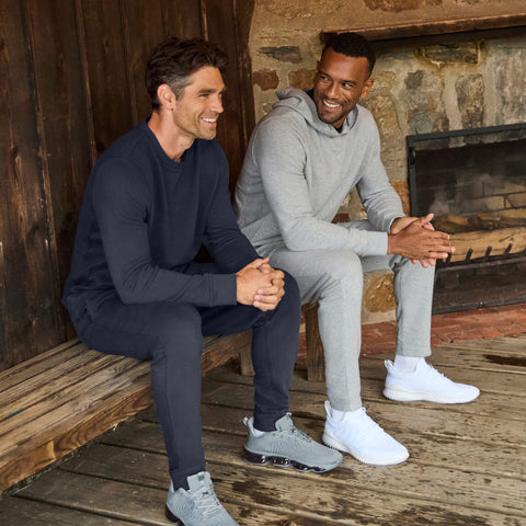 Two men by a stone fireplace, one in a midnight navy crewneck and sweatpants, the other in a gray hoodie and sweatpants.