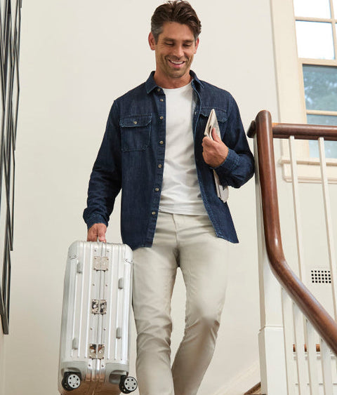 A man walking down a staircase holding a silver hard-shell suitcase and a tablet, wearing a dark blue denim shirt over a white t-shirt and beige pants.