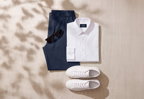 Flat lay of navy trousers, a white dress shirt, sunglasses and white sneakers on linen.