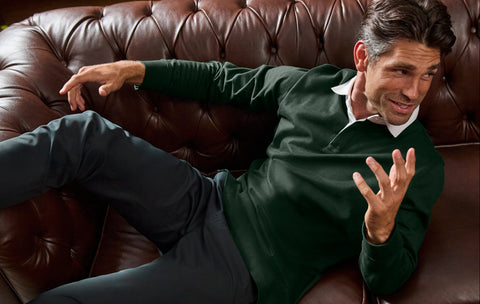 A man reclining on a brown leather tufted sofa, wearing a dark green long-sleeve polo shirt over a white collared shirt and dark trousers.